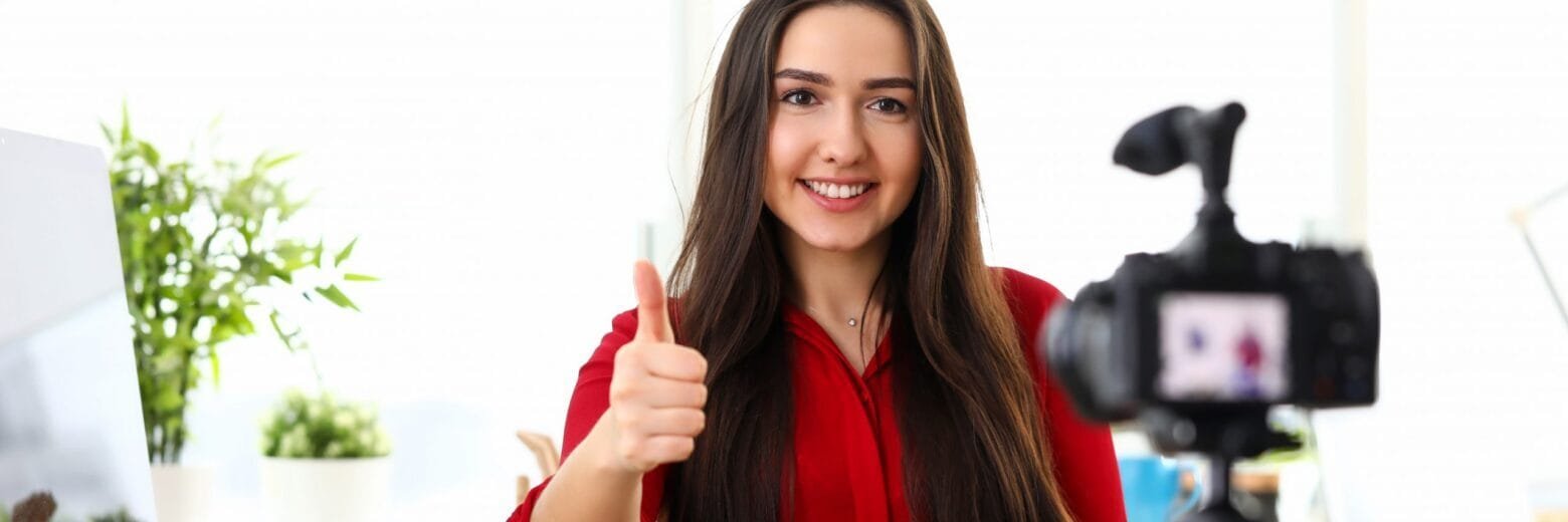 Young woman sitting in office opposite camera and showing good luck thumb finger up