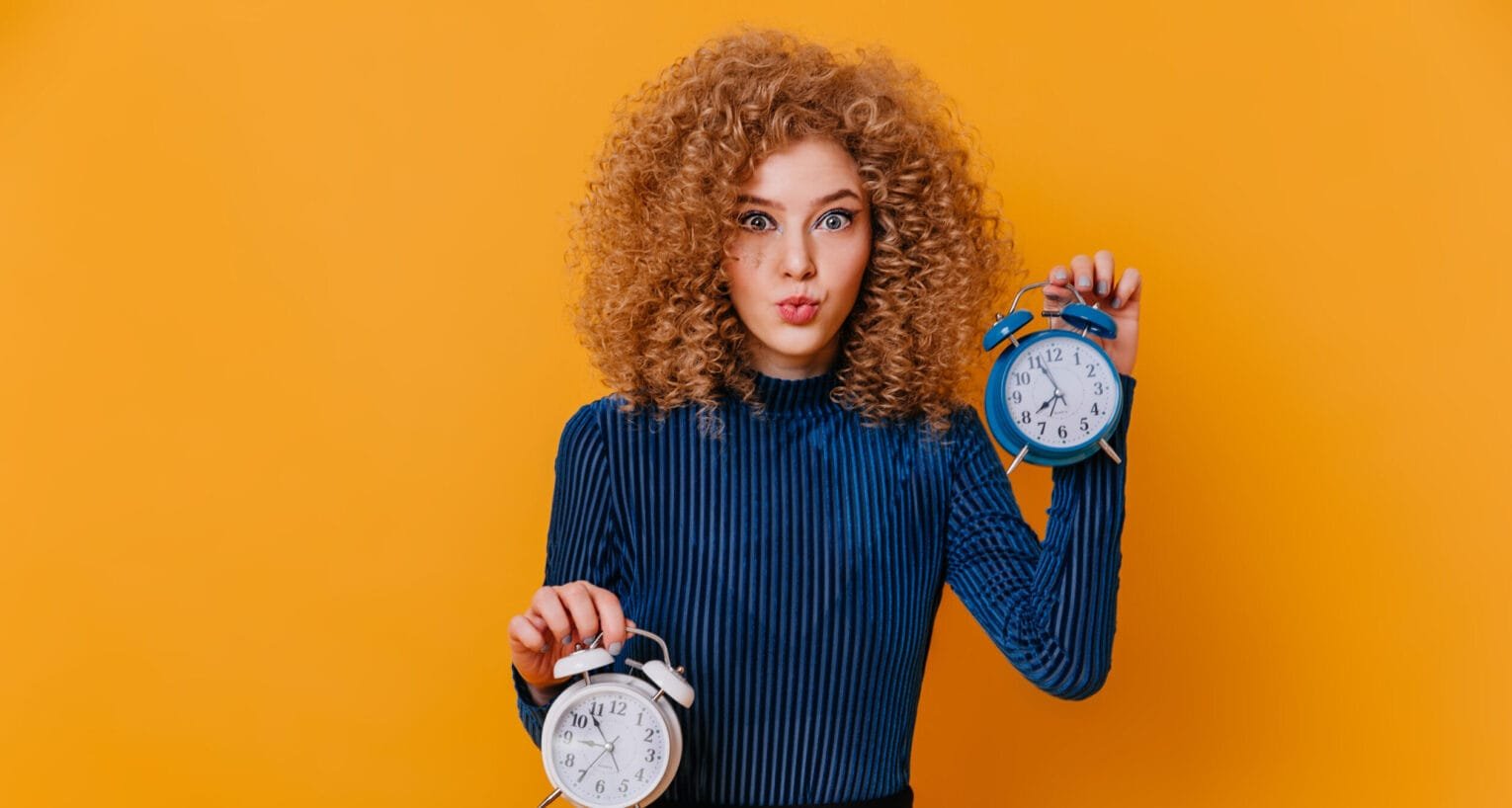 Blue-eyed curly blonde lady in striped sweater and skirt whistles and poses with alarm clocks on yellow background.
